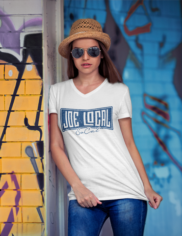 Woman standing against the wall wearing a straw hat and sunglasses. T-shirt shown is white with a blue Joe local so Cal logo. She is wearing blue jeans.