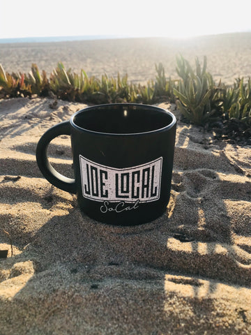 Joe Local 12oz Coffee Mug. Black ceramic mug with white Joe Local So Cal flag logo. Handle is shaped in a half circle allowing for room for your fingers. The mug has a solid  and heavy feel. The mug is round in shape from top to bottom of the mug. 